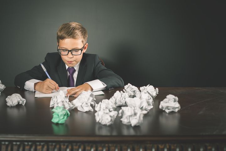 male child in a suit writing a letter, the table next to him covered in crumpled papers